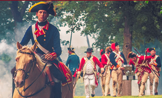 A man in 18th-century military uniform rides a tan horse in the foreground, while several soldiers in red coats march behind him during the Ticonderoga StreetFest in a wooded outdoor setting.